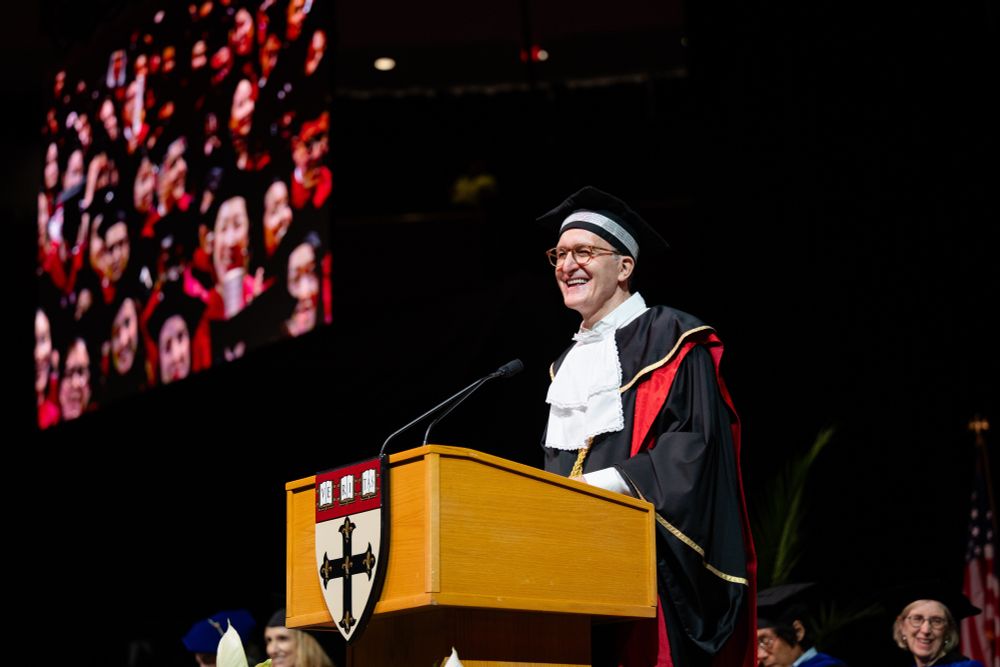 Dean Baccarelli in academic regalia speaks at a podium during the 2025 Convocation ceremony, with a joyful crowd of graduates on a screen in the background. The podium bears the Harvard Chan School emblem.