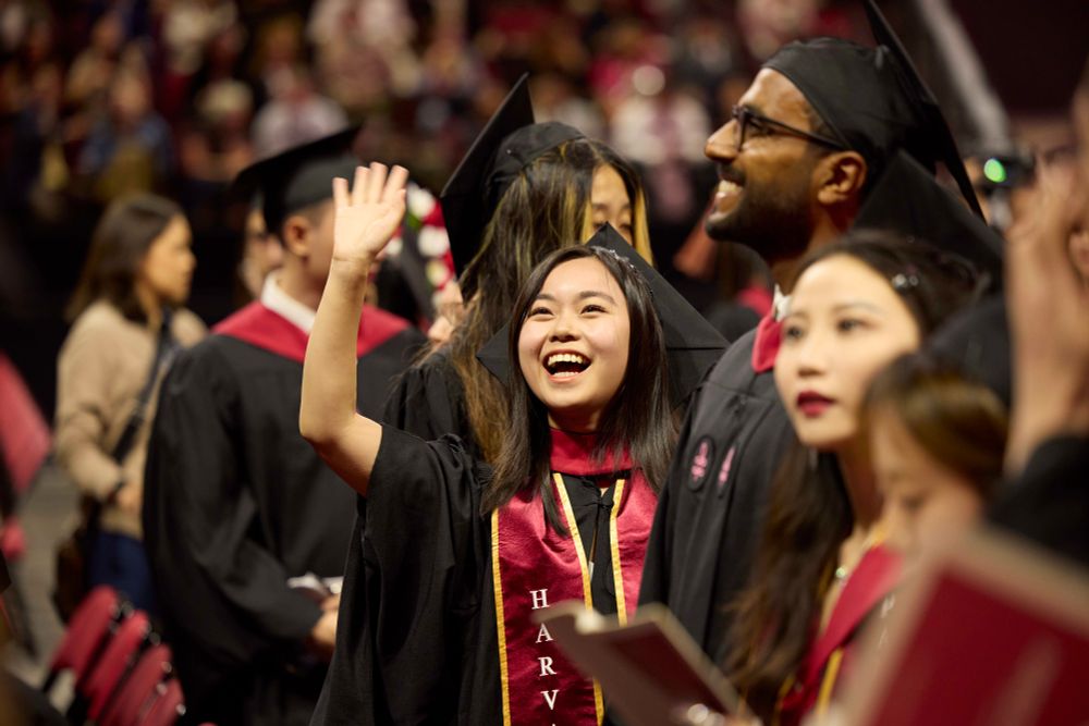 Graduates in caps and gowns at the Harvard Chan School Convocation ceremony, one smiling broadly and waving.