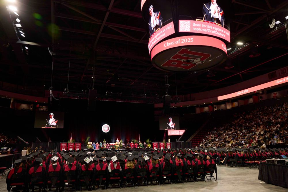 Convocation ceremony at Harvard Chan School with students in caps and gowns seated, listening to Dean Baccarelli visible on large screens in a large arena.
