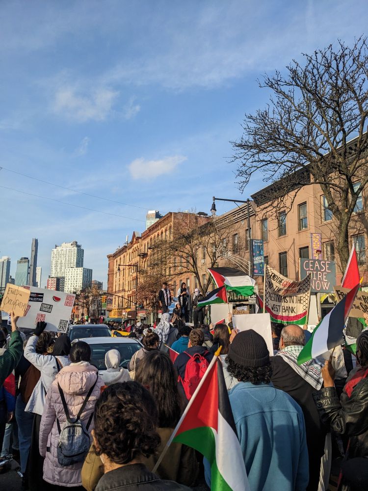 People marching on Flatbush with Palestinian flags and signs calling for ceasefire . Two people wearing keffiyeh stand on top of a truck and face the crowd. Cars are stopped because they can't move through.