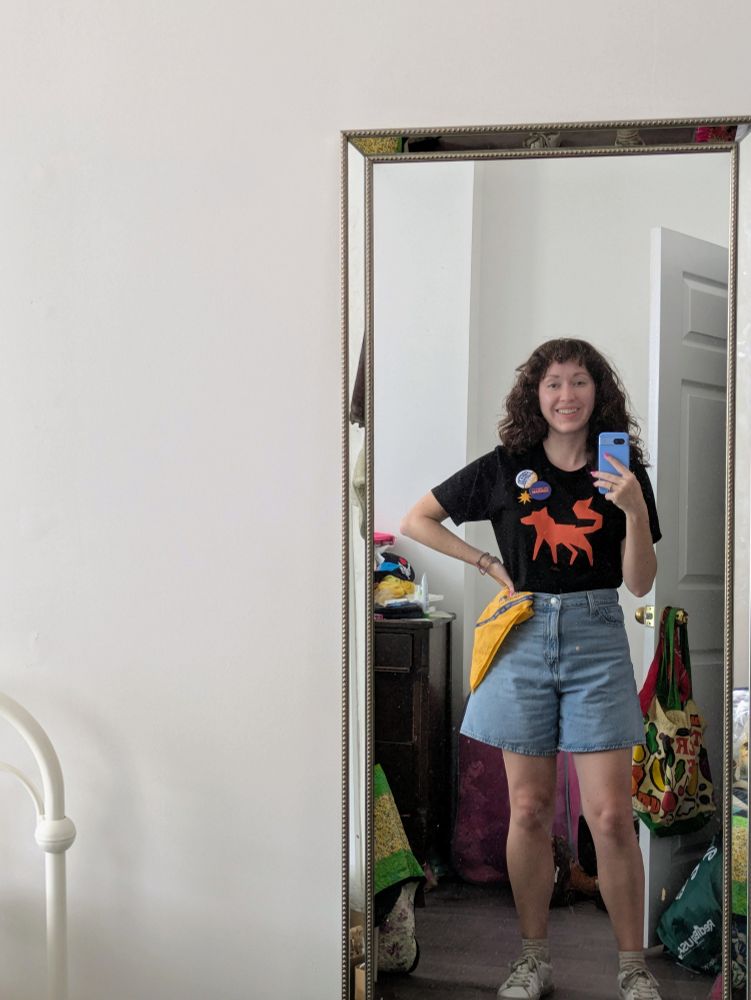 Mirror selfie of Bella. She is smiling and wearing denim shorts and a black tee shirt with the WFP wolf logo. On her shirt she wears "freeze the rent" and "hot girls for Zohran" buttons. On her hip is a yellow Zohran for Mayor bandana. 