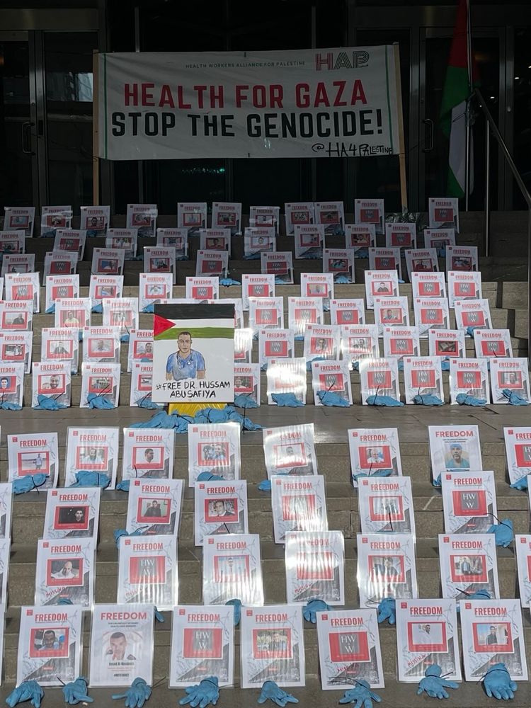 The steps to the Israeli consulate at 2 Bloor St East in Toronto. Placards are arranged on the steps. Each has the name and photo of a health worker held currently in Israeli detention. There is a blue surgical glove tied with a zip tie, in front of each picture.