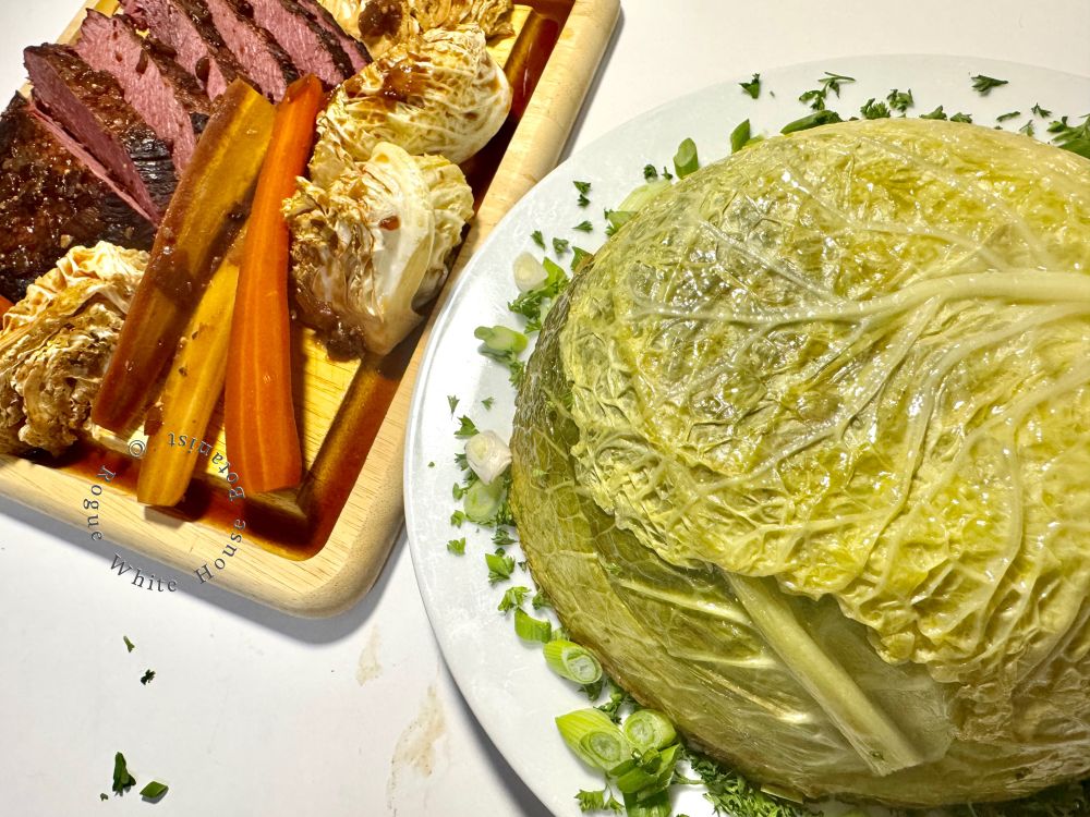 Charlotte de Chou (French stuffed cabbage) on a white plate, illustrating the lacy veins of a savoy cabbage shown adjacent to sliced corned beef, rainbow carrots, and chunks of cabbage on a wooden serving board. 