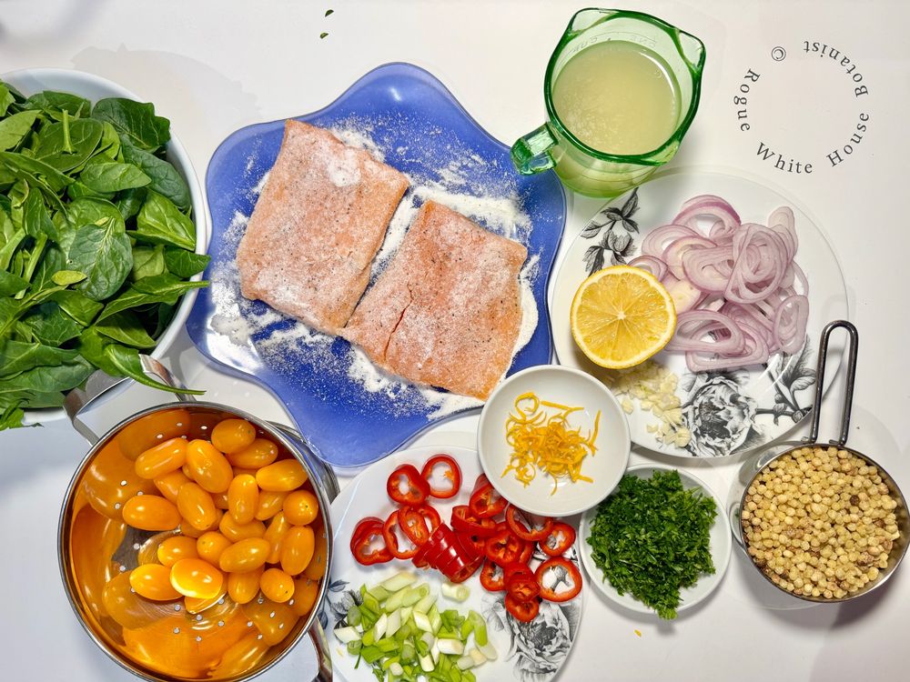 All the ingredients for Wild Mountain Trout with Sauteed Spinach & Veggies served over Fregola Sarda. Clockwise from top left: a bowl of spinach, wild trout dusted with rice flour, wine &chicken stock in a green measuring cup, a plate with lemon, shallots and garlic, a cup full of uncooked Fregola Sarda, some parsley, lemon zest, red baby bell slices and green onion, and a bowl of yellow tomatoes. 