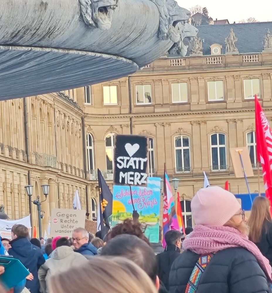 Im Hintergrund ist das Stuttgarter Neue Schloss zu sehen. Ganz links ragt der Südliche Springbrunnen ins Bild. Vor dem Schloss sind viele Demonstrierende zu sehen sowie mehrere Transparente und Plakate mit Aufschriften wie "Herz statt Merz", "Schöner leben ohne Nazis", "Gegen rechte Unterwanderung". Außerdem einige Fahnen, darunter eine Transgender-Flag.