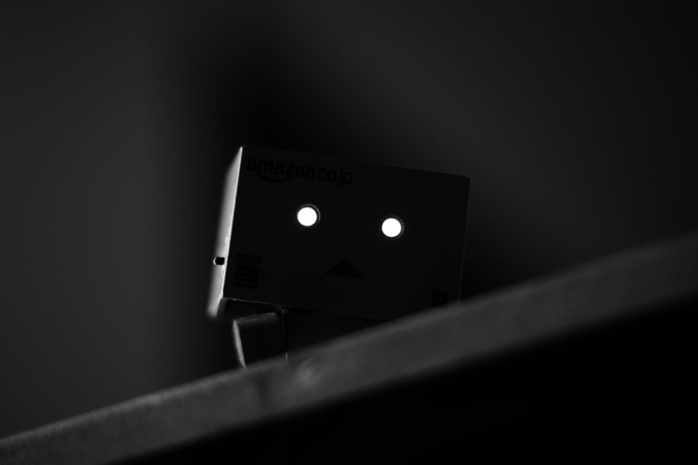 A monochromatic photograph of a fictional cardboard box robot character figurine called Danbo, with the eyes turned on, looking down from a coffee table, in a supposedly spooky manner.