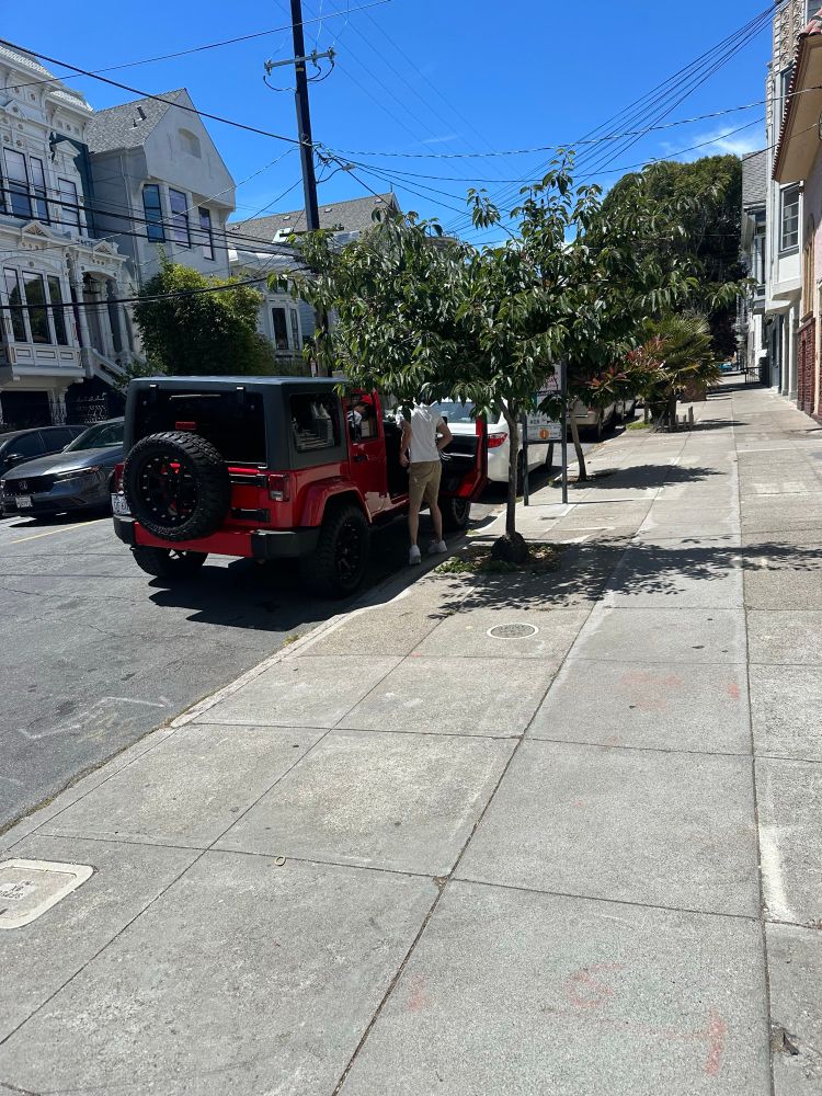 Red Jeep with a partially visible license plate reading 'DF 60,' and a man in khaki shorts and a white shirt standing to the right of it, positioned behind the front door—apparently to conceal that he had just urinated in plain view during daylight