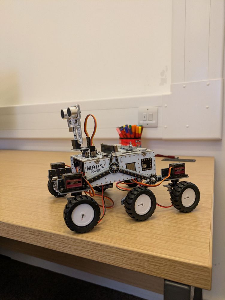 A photo of a mini Mars Rover model on a wooden desk.