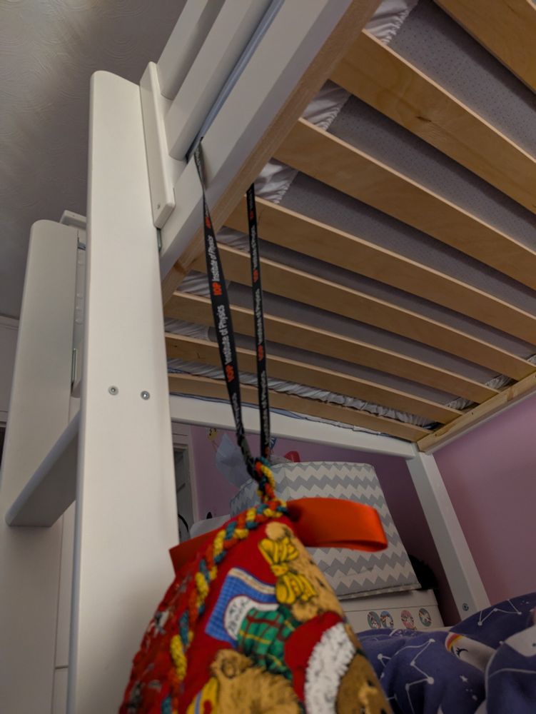 A photo taken from the floor looking up at the underneath of a top bunk bunk bed. There's a black Institute of Physics lanyard looped around the railing with a colourful Christmas stocking attached to the other end. 