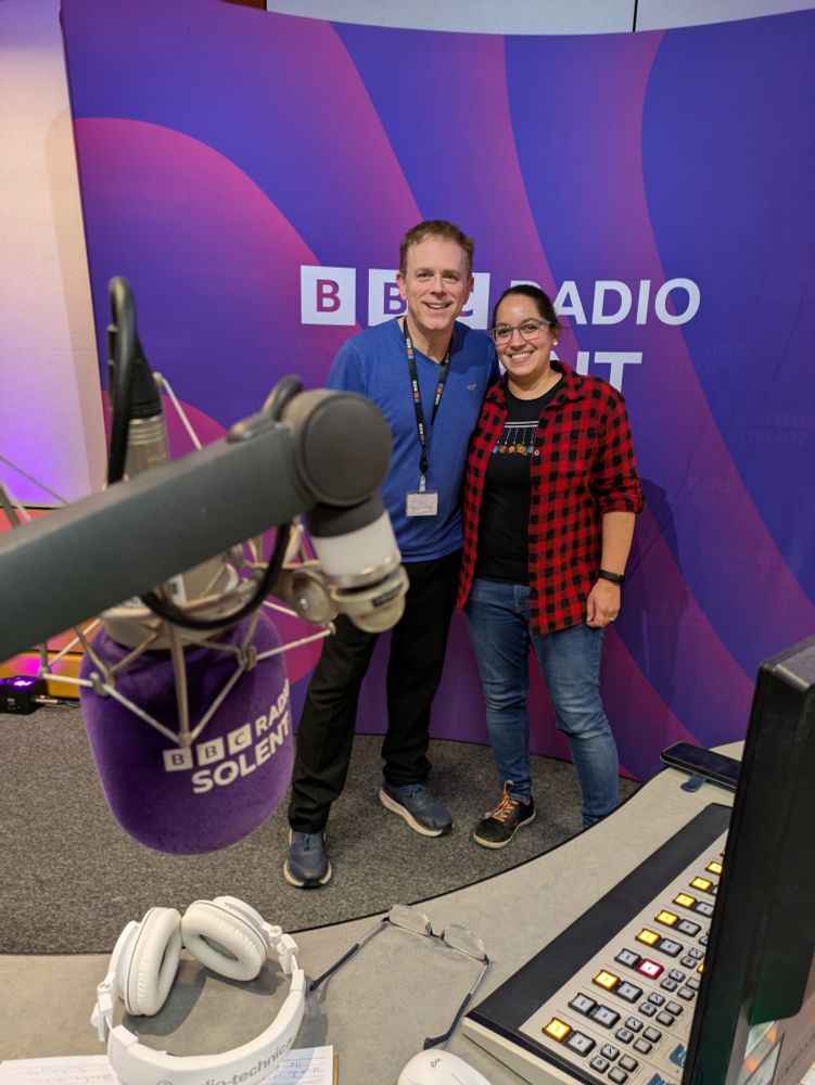 A photo of me in the radio studio with Chris Jarvis the presenter. We are standing next to each other in front of a BBC Radio Solent background and smiling at the camera. Chris is on the left of the photo in a blue t-shirt and black trousers. I'm on the right of the photo wearing a red and black check shirt over a black t-shirt and blue jeans. The radio desk is in the foreground of the photo with a purple BBC Radio Solent microphone hanging down.