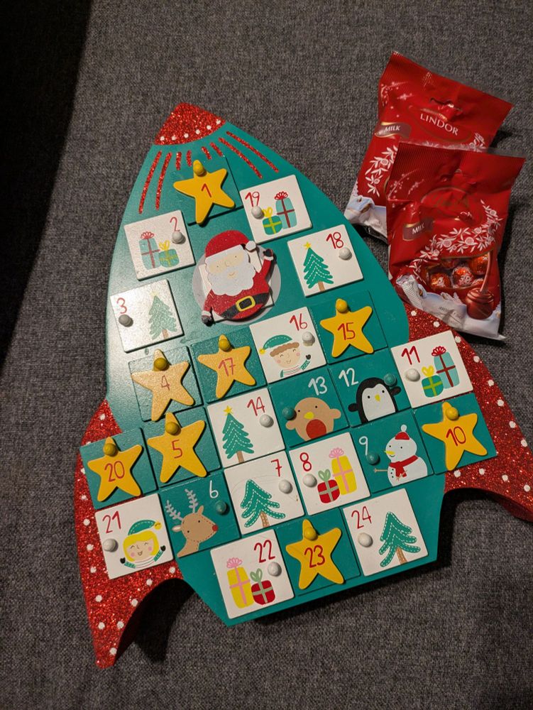 A photo looking down at a wooden advent calendar in the shape of a rocket, with two packets of small chocolates next to it on the right. The rocket is green with a glittery red top and fins. Most of the rocket has drawers on it which are either green or white with Christmas designs on them.