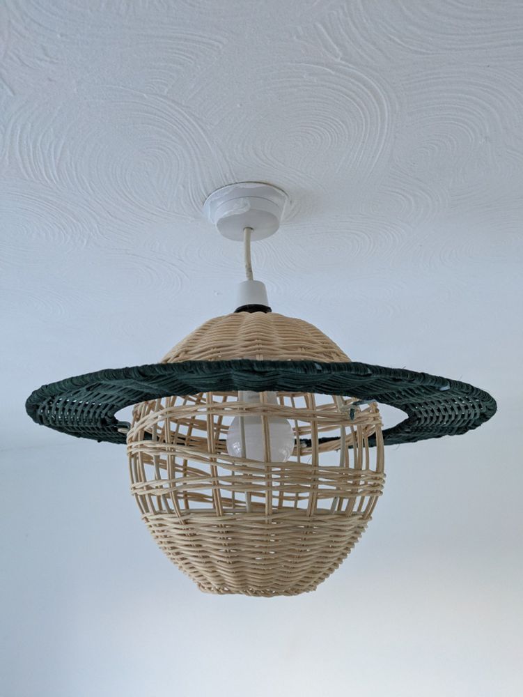 A photo of a Saturn shaped lampshade hanging from the ceiling. It's made from rattan. The sphere of the planet is a natural light brown wood colour and the ring is a dark green/black colour.