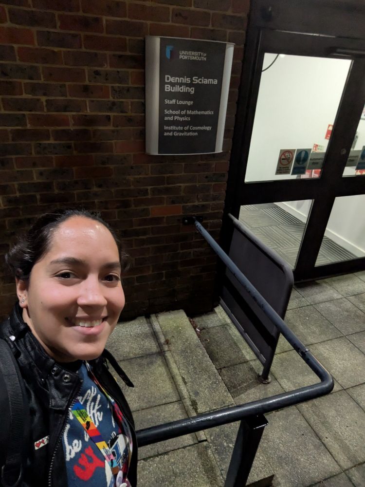 A selfie of me outside my office building. I'm to the left of the photo wearing a Christmas jumper with a black jacket over it. You can see the building behind me with a sign that says "Dennis Sciama Building" and "Institute of Cosmology and Gravitation".