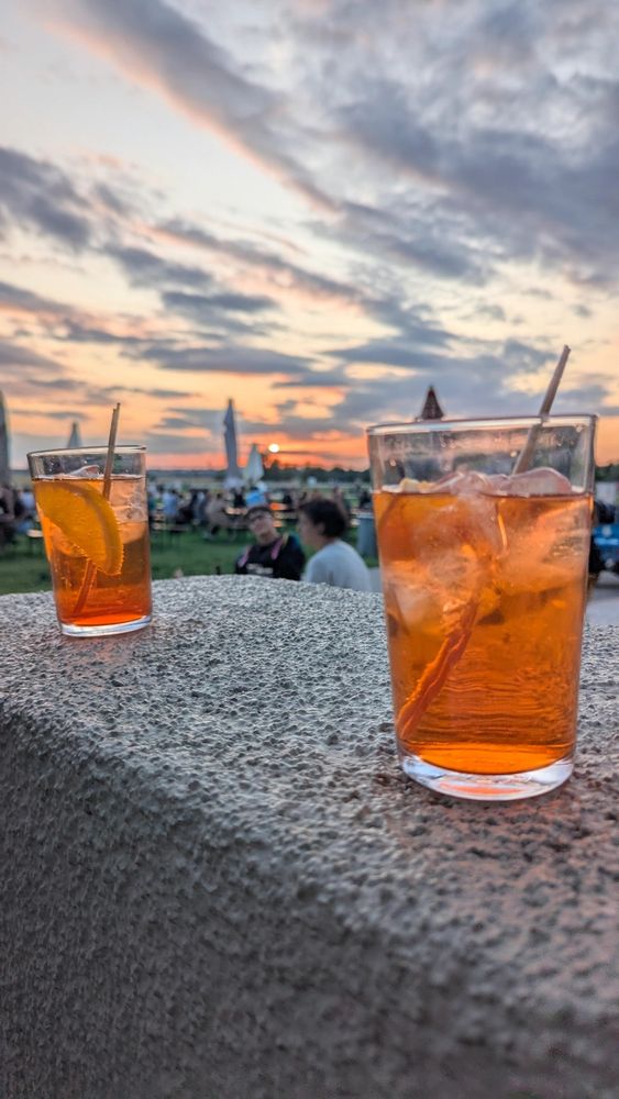 Zwei Gläser mit Aperol Spritz gefüllt stehen auf einer Mauer. Im Hintergrund ein leicht wolkiger Himmel bei Sonnenuntergang und eine Wiese voller Menschen.