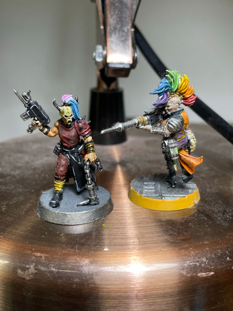 A pair of female Warhammer models. One on the left is in red armour, carrying a pistol and a chain and in wearing a skull mask. Her hair is blue, white and pink. On the right is an Escher hanger with a needle rifle, with orange and purple clothing. She has rainbow hair