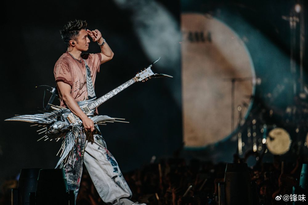 Nicholas Tse holding a guitar shaped like the head/torso/wings of an angel, while saluting, who knows, probably his band 