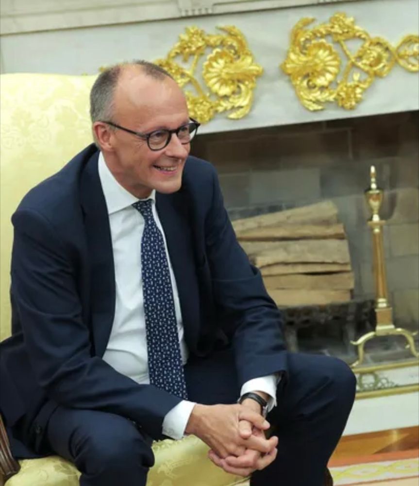 Merz sitzt grinsend auf einem stuhl im weißen haus 
