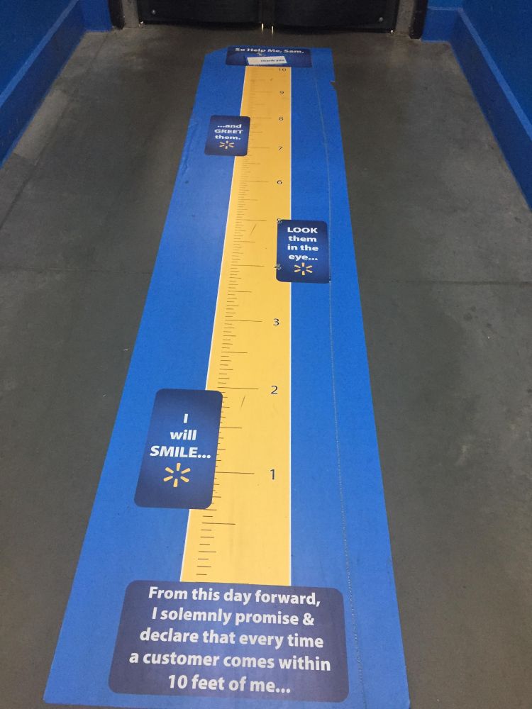 A 10 foot long blue floor decal with Walmart branding reading "From this day forward, I solemnly promise & declare that every time a customer comes within 10 feet of me... I will SMILE... LOOK them in the eye... and GREET them. So Help Me, Sam."