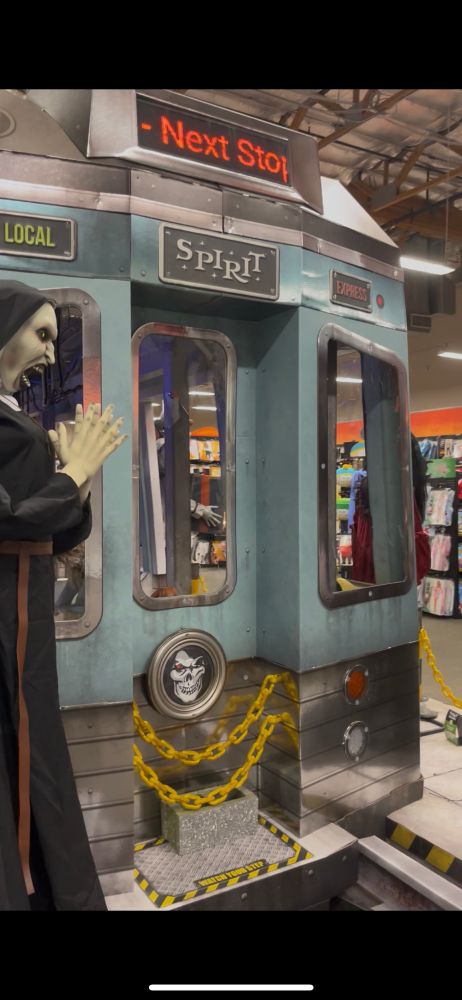 front of a creepy cardboard “subway” train in spirit halloween with skulls and a haunted nun and shit