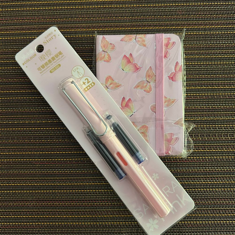 A small pink notebook with
Butterflies on the cover displayed with a pink fountain pen and two cartridges, still in the packaging. 