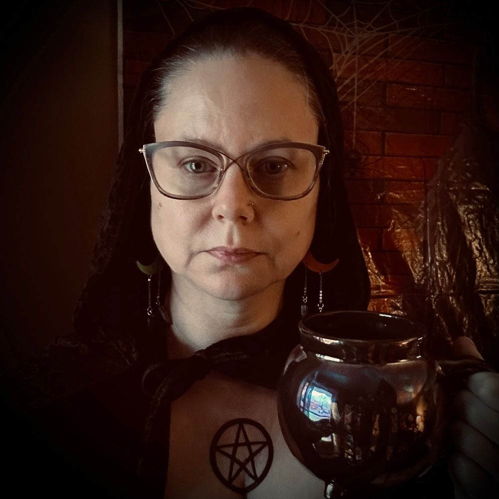 A woman wearing a hooded black cloak and gold-frame glasses looks directly at the camera. There is a large black pentagram temporary tattoo on her upper chest.   She holds a black mug shaped like a cauldron.  