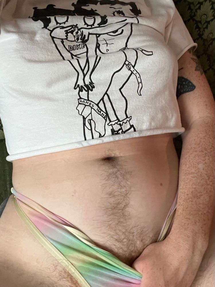 dayze, white tattooed and freckled person, sits on a green couch and pulls down their panties to show off their happy trail. they are wearing white crop top with two betty boops on it. 