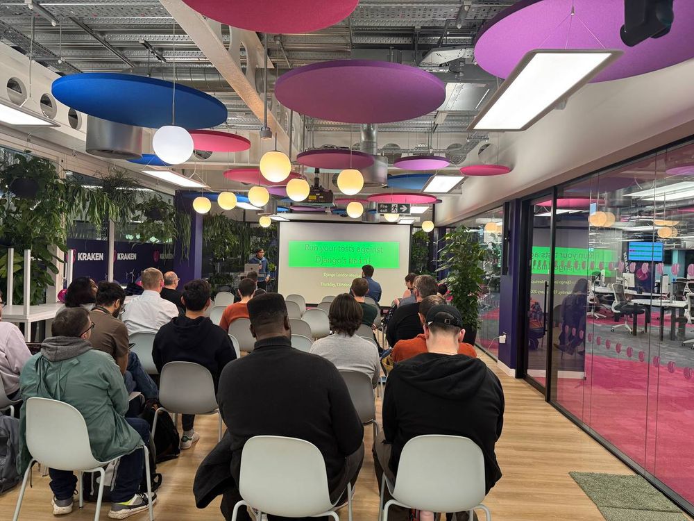 A meetup event for Django London is taking place in the Kraken Tech event space. Attendees are seated in rows of white chairs, facing a speaker at the front of the room. The speaker, Sage Abdullah, is presenting a talk titled "Run your tests against Django's main!" A projector screen displays the presentation title in green text. The modern event space features colorful circular ceiling panels, hanging lights, glass walls, and indoor plants. In the background, an open office area with desks and monitors is visible through the glass partition.



