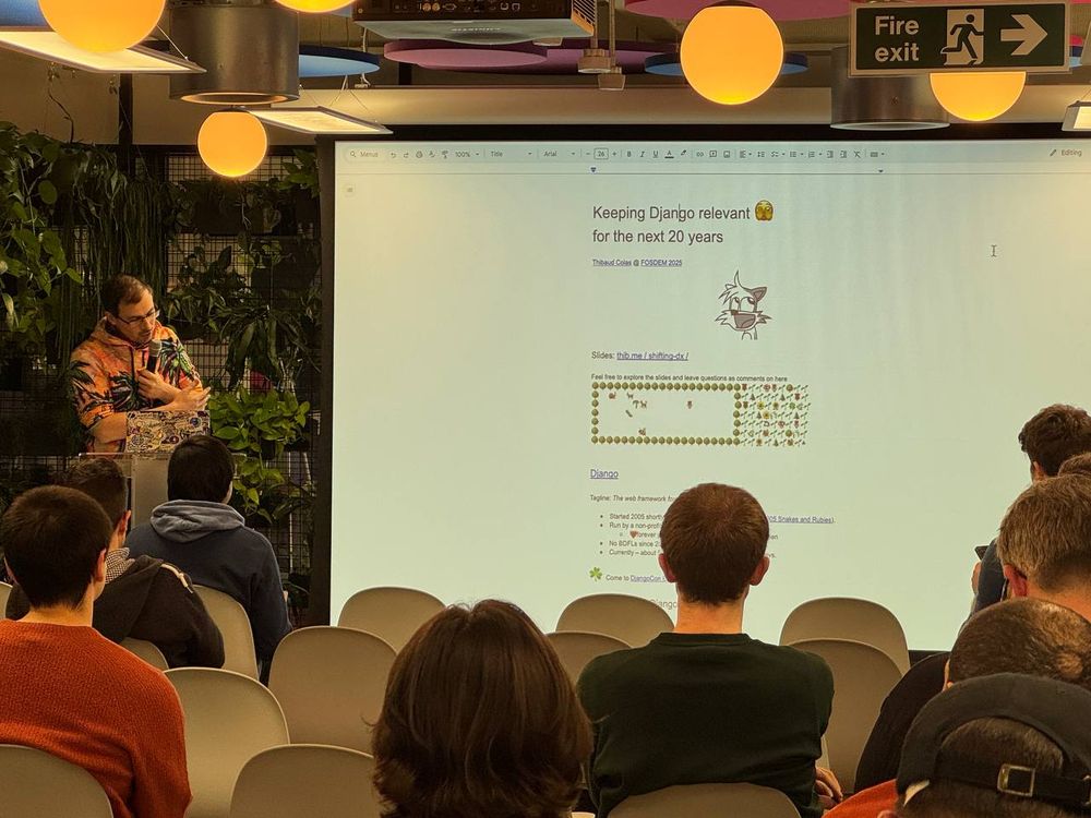 A Django London meetup is taking place at the Kraken Tech event space. The second speaker, Thibaud Colas, is presenting a talk titled "Keeping Django relevant for the next 20 years." He stands at a transparent podium, gesturing towards a large projector screen displaying his google doc presentation. The slide features the talk’s title, a cat illustration, and a diagram. The audience, seated in rows of white chairs, listens attentively. The room is decorated with indoor plants, colorful ceiling panels, and hanging lights, creating a modern and vibrant atmosphere.
