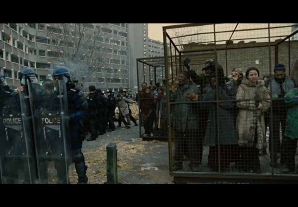 Frame from the 2006 movie "Children of Men." Under gray city skies, illegal immigrants are held in open-air cages while riot police hold off protesters.