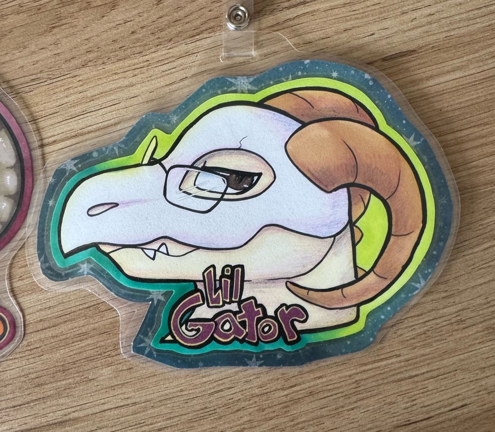 Badge from 2021 by Candy (@plsfeedthebear.bsky.social) that’s been laminated. It reads “Lil Gator”, which was the name of my sona at the time.

It’s a side profile of an alligator with horns wearing a helmet made of bone and a pair of rectangular glasses; his visible eye his half closed with a smirk or smile.