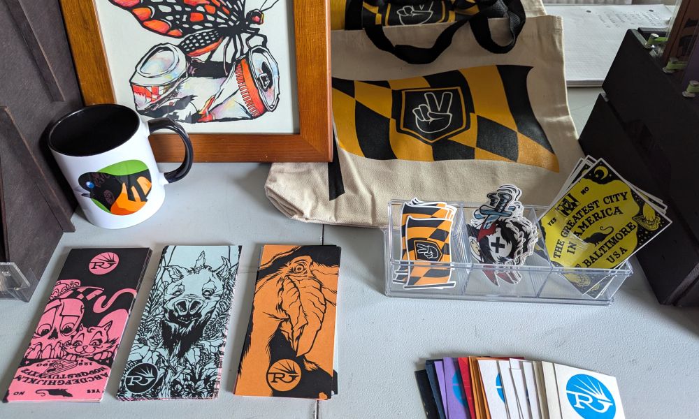 Top view of the center of my staged market table including a framed hand finished print, screen printed bookmarks, stickers, an example tote bag, the mints cup and business cards.  