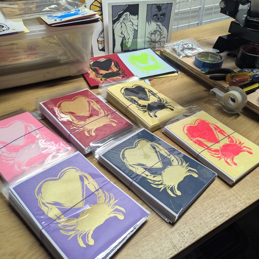 Screen printing table in my basement. In the foreground are stacks of finished cards with a crab holding a heart design in 6 color combinations. Cards are printed by me on a variety of color card stock from French paper co.