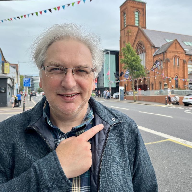 Mé seanding on the street pointing over my shoulder at a red brick building behind me.