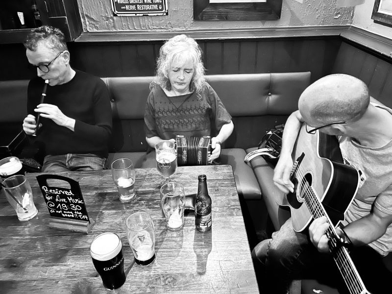 A man playing tin whistle, a woman playing concertina and a man playing guitar at a table in the corner of a pub.