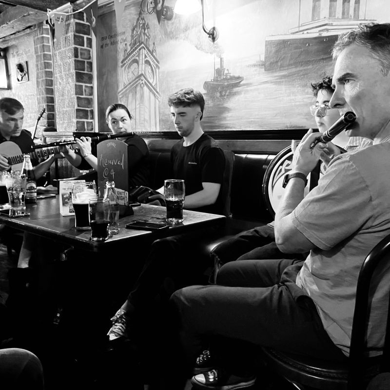 A flute player in the foreground with bodhrán, concertina, flute and guitar in the other side of the table.