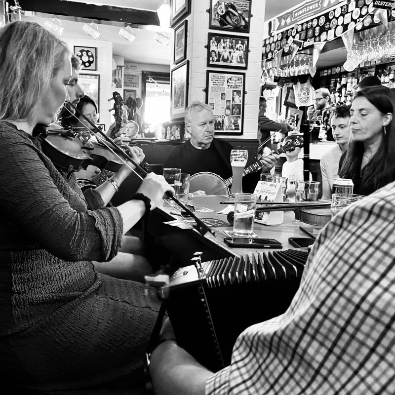A woman playing fiddle, a man playing banjo and another man playing button box round a pub table.