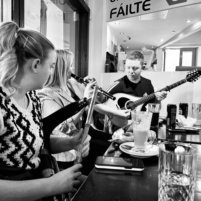 A woman playing pipes, a woman playing flute, and a man playing bouzouki at a café table under a sign saying “fáilte.”