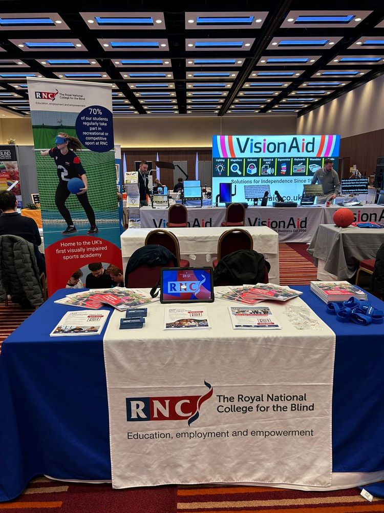 The RNC exhibition stand with prospectuses, flyers, signature guides, wristbands, and a branded tablecloth. A single banner stands beside the table, highlighting how RNC supports visually impaired students through sport