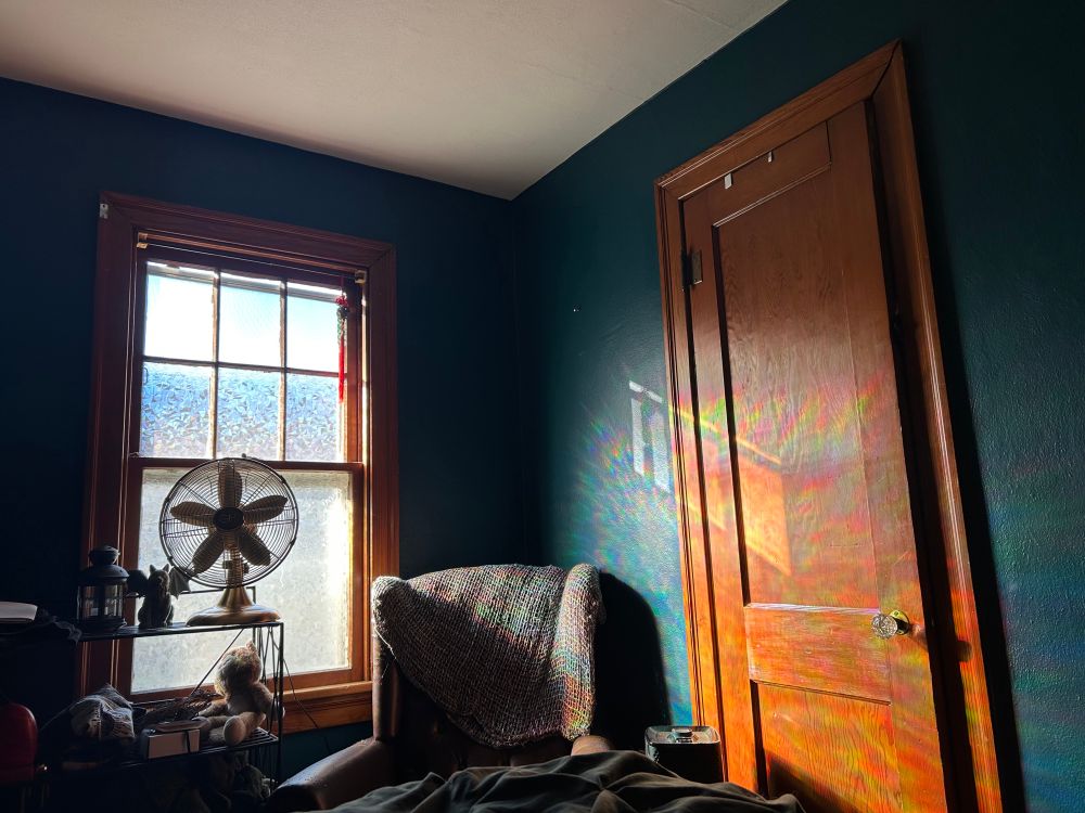 Rainbow beams from window cover in a wooden door with a chair and a gold metal fan on a shelf.