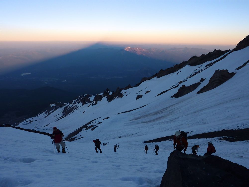 A picture from the side of Mount Shasta, some climbers in the foreground and the huge triangular shadow of the mountain extending to the horizon. 