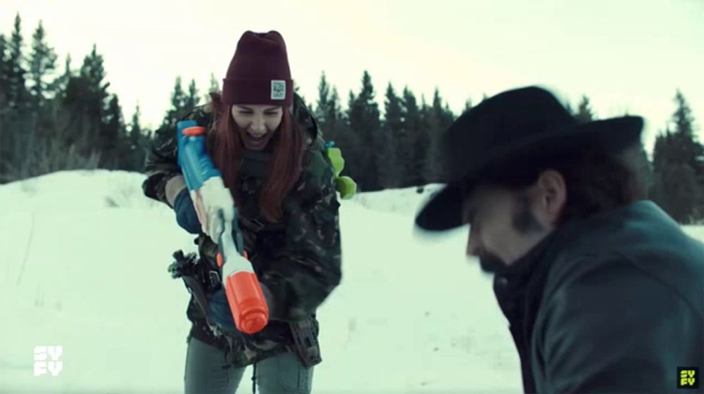 a man and a woman are playing with water guns in the snow and the woman is wearing a beanie that says no