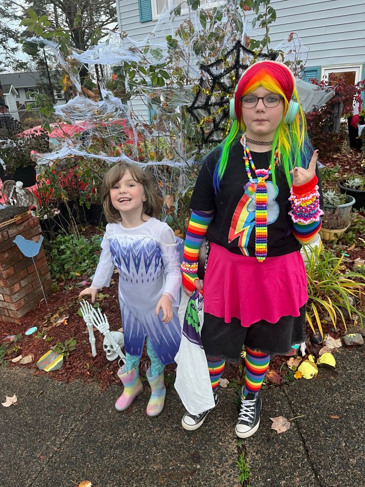Small human dressed in an Elsa dress and a larger small human cosplaying a My Little Pony in a rainbow wig
