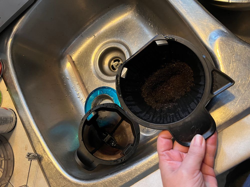 A clean reusable coffee filter in a silver sink next to a black coffee maker basket full of used coffee grounds. 