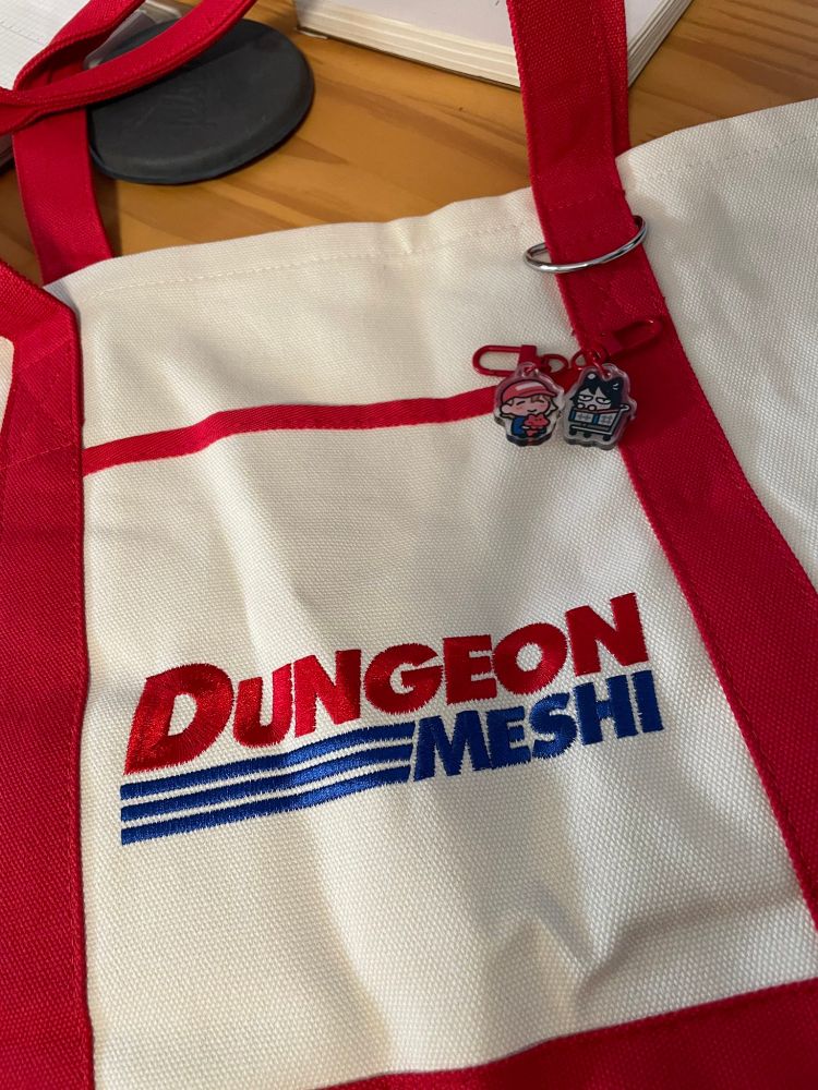 a photo of a sturdy tote bag with a stylized logo that reads "Dungeon Meshi", but in the costco style. The bag is white and red. There are very small and cute acrylic charms of a chibi falin and a chibi izutsumi on the bag.