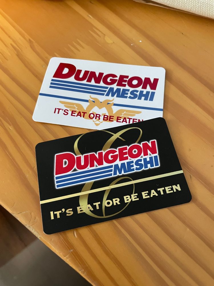 a photo of two cards that say "Dungeon Meshi: it's eat or be eaten", styled like Costco membership cards. 