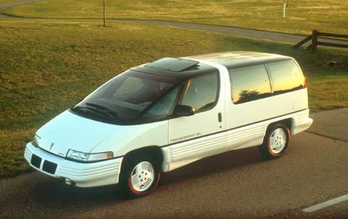 Image of a white sleek and sci fi looking mini van on a road with a grassy field in the back ground. This is the long gone 1991 Pontiac Trans Port.