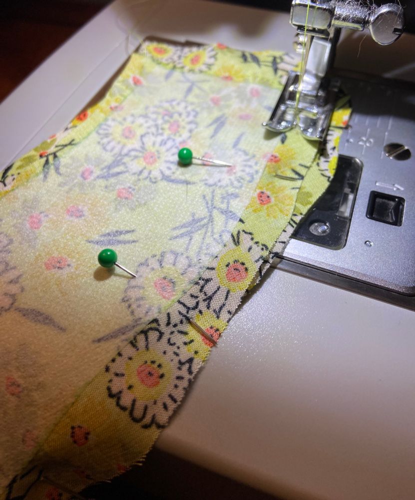 pinned together fabric in the midst of being sewn together on a sewing machine.  the fabric is a yellowy-green color with white flowers that have a yellow and orange center.  most of the designs are outlined in black.  in a few places there are also some green flowers either orange centers. 