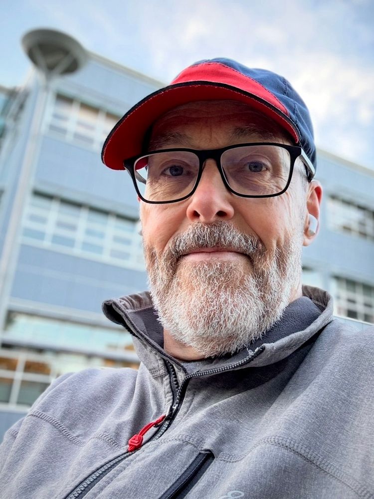 A middle-aged man is sitting on a bench outside of a university library. He’s wearing black-rimmed glasses, a red and blue cap and a grey jacket. 