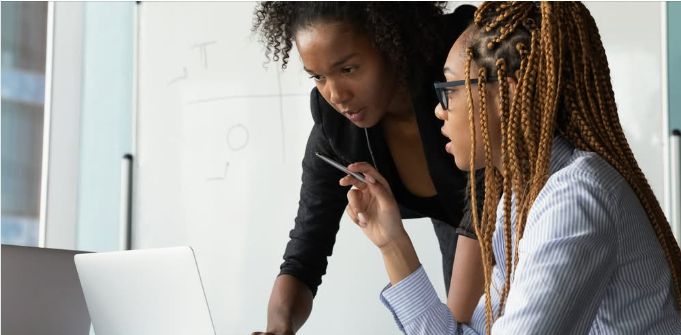 Two co-workers look at a laptop in front of a whiteboard.