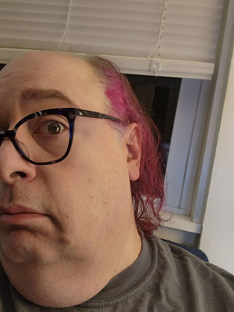 A close-up of the author. You can see the left side of their face, and their newly dyed pink hair. Also, tortoiseshell cat's eye glasses.